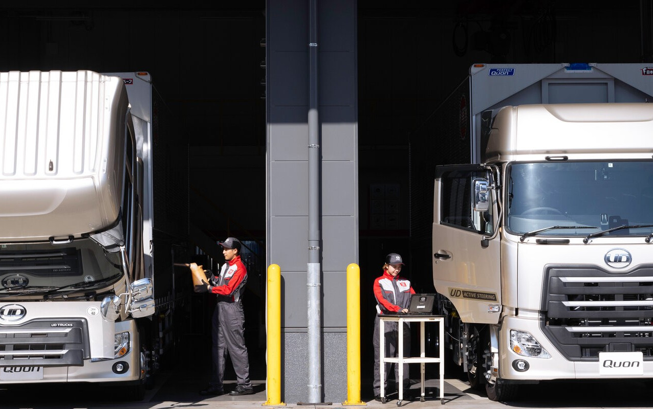 Two workers in red uniforms check clipboards beside parked delivery trucks.