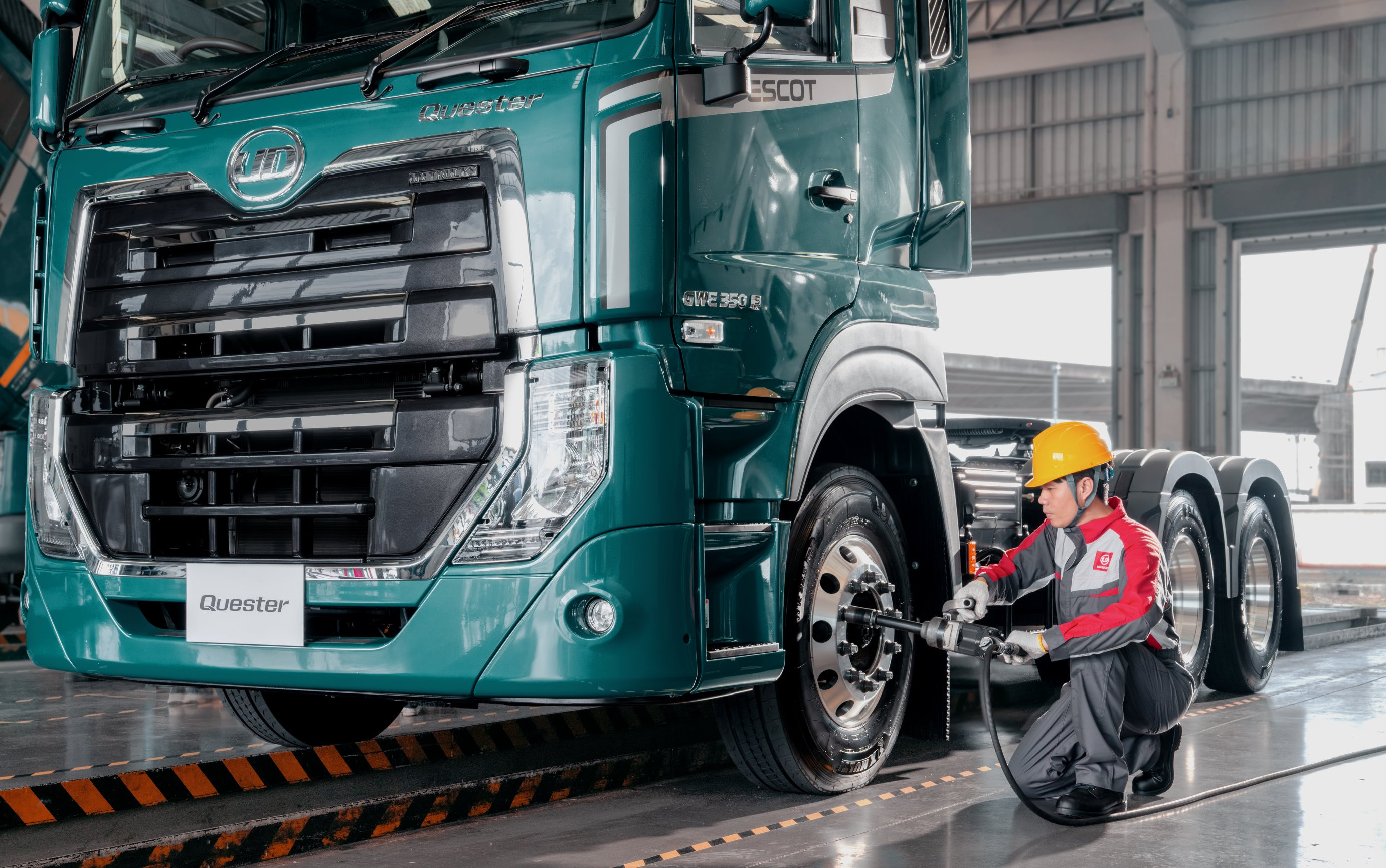 Mfanyakazi amevaa kofia ya njano akikagua tairi la lori kubwa kwenye gereji.