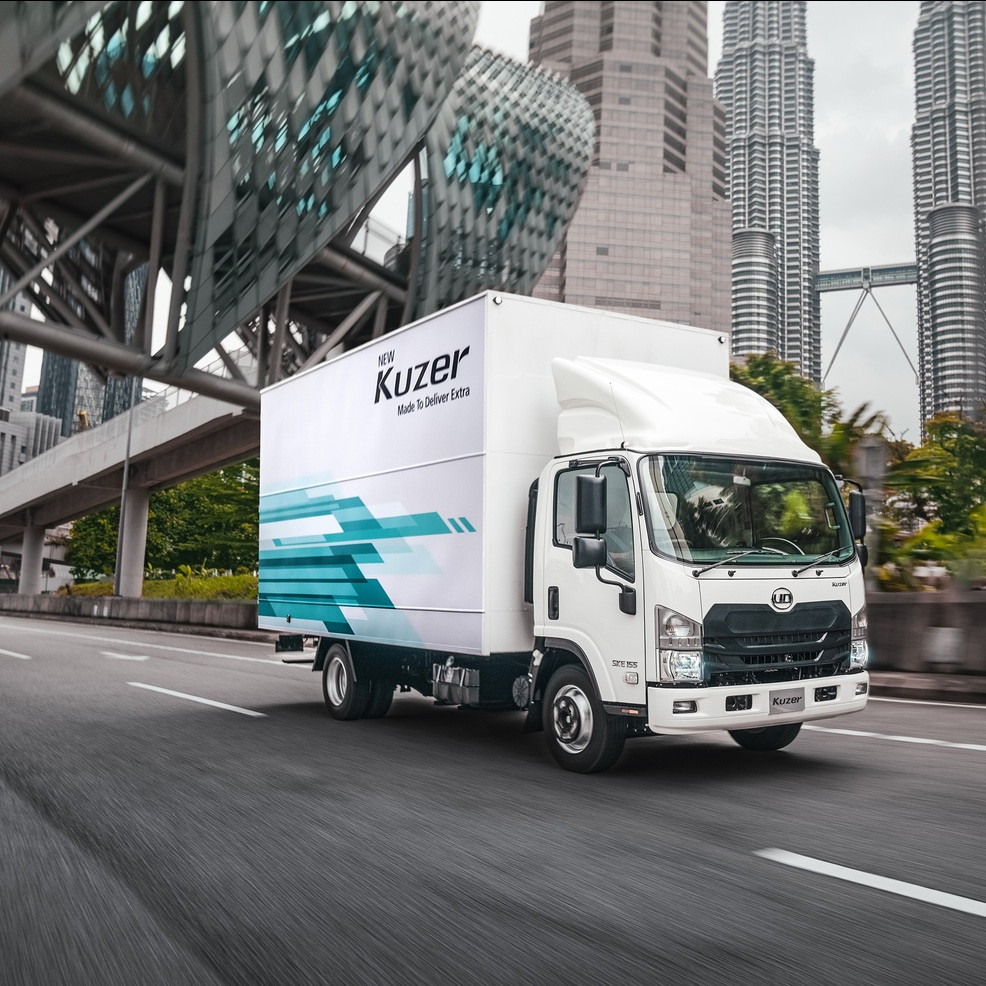 White delivery truck driving on a city street with tall buildings in the background.