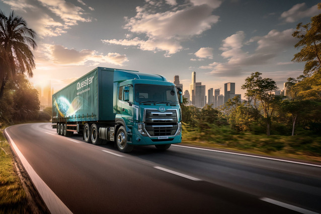 Blue semi-truck driving on a highway with a city skyline in the background at sunset.