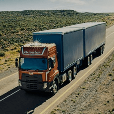 Truk kontainer biru melaju di jalan raya sepi, dikelilingi pemandangan alam.