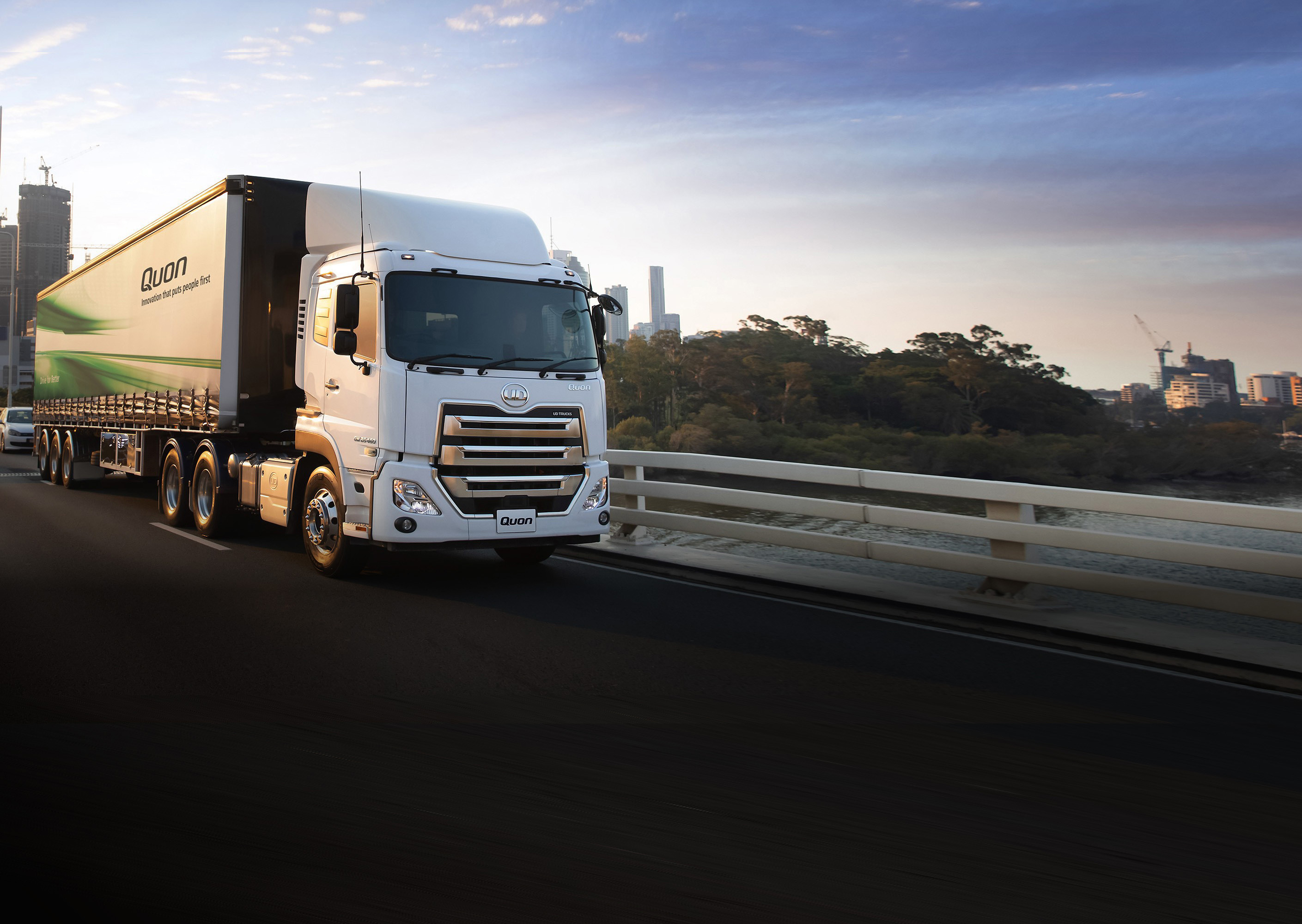 White semi-trailer truck driving across a bridge at sunrise, city skyline in background.