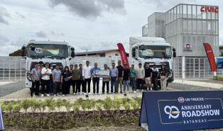 Grupo ng tao sa harap ng dalawang trak, may gusali at event banner sa likod.