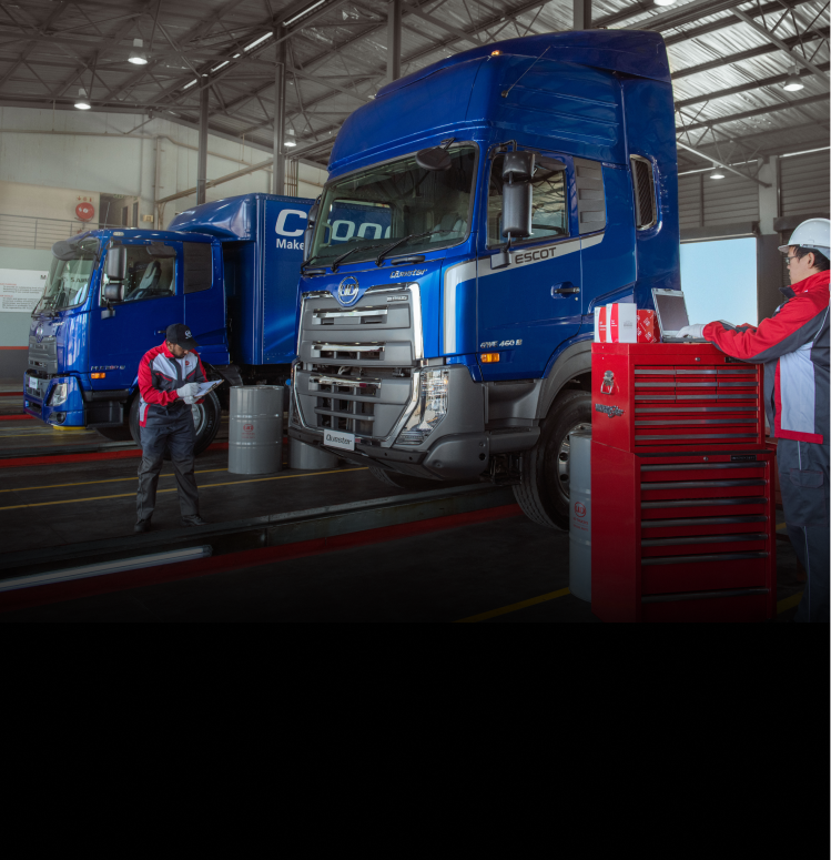 Two mechanics work on blue trucks in a brightly lit service garage.