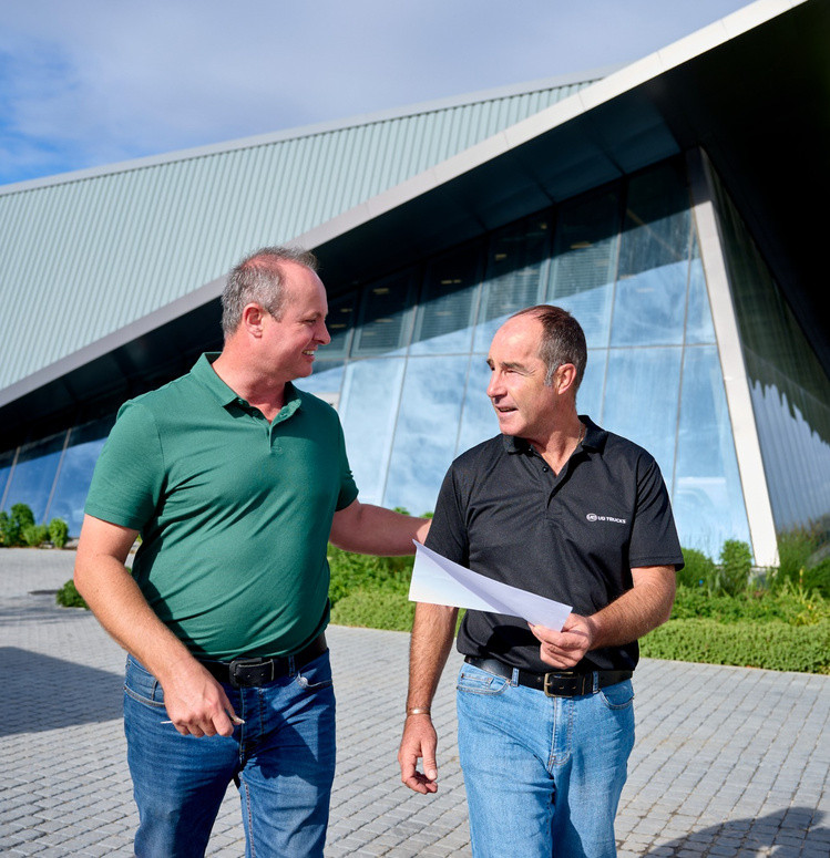 Two men talking and walking outside a modern glass building on a sunny day.